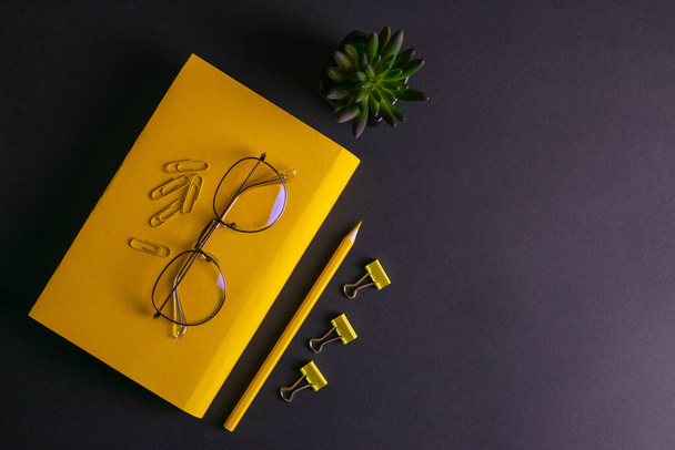 Gelber Bleistift, gelbes Notizbuch und Brille, Stecknadeln, Klemmpapier auf schwarzem Hintergrund. - Foto, Bild