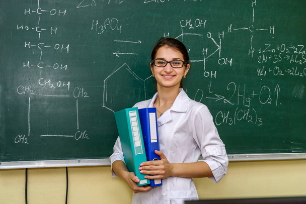 Chemist with folders standing and posing for camera. Behind woman university board with chemical formulas - Photo, Image
