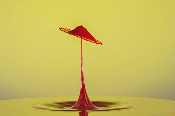 Red water droplets colliding to form an umbrella shape against a yellow background - Photo, Image