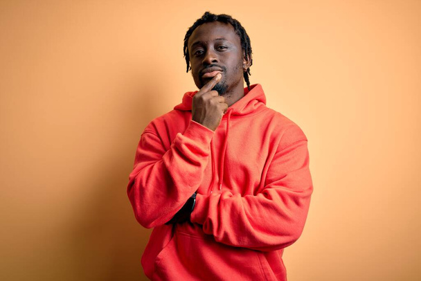 Young african american sporty man wearing sweatshirt with hoodie over yellow background looking confident at the camera with smile with crossed arms and hand raised on chin. Thinking positive. - Φωτογραφία, εικόνα