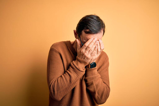 Hombre alzado de mediana edad que usa suéter marrón y gafas sobre un fondo amarillo aislado con expresión triste que cubre la cara con las manos mientras llora. Concepto de depresión
. - Foto, imagen