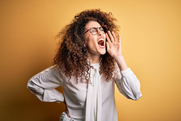 Young beautiful brunette woman with curly hair and piercing wearing shirt and glasses shouting and screaming loud to side with hand on mouth. Communication concept. - Photo, Image