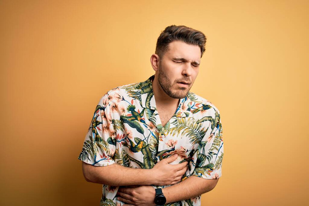 Young man with blue eyes on vacation wearing floral summer shirt over yellow background with hand on stomach because nausea, painful disease feeling unwell. Ache concept. - Фото, изображение