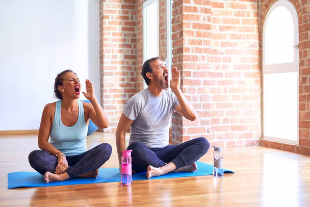 Middle age sporty couple sitting on mat doing stretching yoga exercise at gym shouting and screaming loud to side with hand on mouth. Communication concept. - Photo, Image