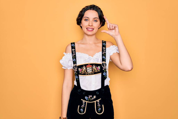 青い目をした若い美しいドイツ人女性は、伝統的な10月祭のドレスを着て笑顔と指を見て、カメラと小さなサイズの看板を行う手で自信を持ってジェスチャー。概念の測定. - 写真・画像
