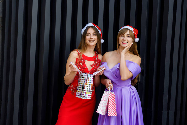 Dvě úžasné ženy v Santa klobouky a elegantní večerní šaty drží nákupní tašky - Fotografie, Obrázek