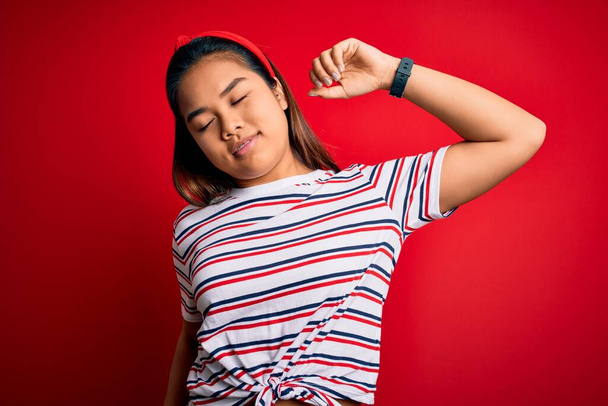 Joven hermosa chica asiática vistiendo casual rayas camiseta sobre aislado rojo fondo estiramiento espalda, cansado y relajado, somnoliento y bostezar para la madrugada
 - Foto, imagen