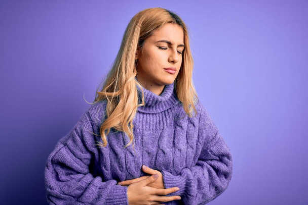 Young beautiful blonde woman wearing casual turtleneck sweater over purple background with hand on stomach because indigestion, painful illness feeling unwell. Ache concept. - Фото, изображение