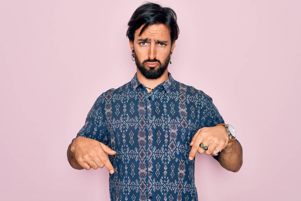 Young handsome hispanic bohemian man wearing hippie style over pink background Pointing down looking sad and upset, indicating direction with fingers, unhappy and depressed. - Photo, Image