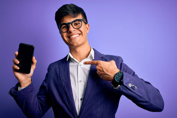 Fiatal jóképű üzletember visel kabát és szemüveg gazdaság smartphone mutatja képernyő meglepett arc mutató ujját magát - Fotó, kép