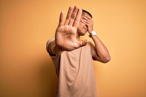 Młody, przystojny, afrykański mężczyzna ubrany w luźny t-shirt, stojący nad żółtym tłem, przykrywający dłońmi oczy i wykonujący stop gestem smutku i strachu. Zawstydzona i negatywna koncepcja. - Zdjęcie, obraz