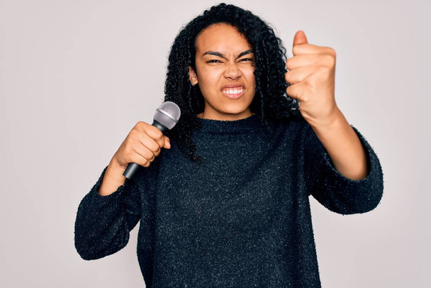 Молода афроамериканка кучерява співачка співає з мікрофоном на білому фоні роздратована і розчарована криками з гнівом, божевільною і криком з піднятою рукою, гнівною концепцією - Фото, зображення