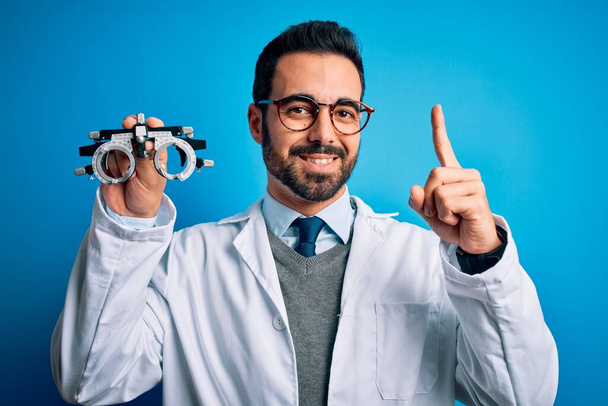 Young handsome optical man with beard holding optometry glasses over blue background pointing finger up with successful idea. Exited and happy. Number one. - Foto, imagen