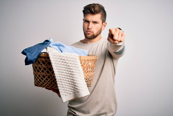 Young blond man with beard and blue eyes doing chores holding wicker basket with clothes pointing with finger to the camera and to you, hand sign, positive and confident gesture from the front - Photo, Image