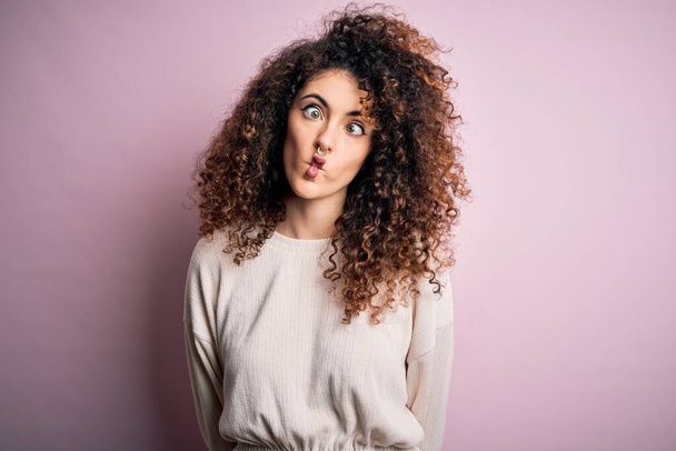 Beautiful woman with curly hair and piercing wearing casual sweater over pink background making fish face with lips, crazy and comical gesture. Funny expression. - Fotografie, Obrázek