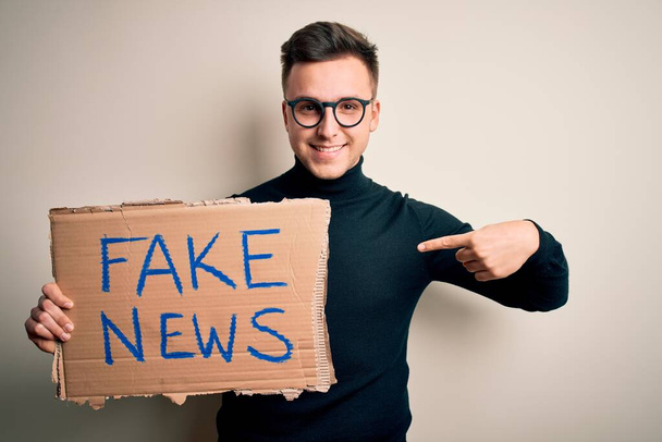 Молодий красивий кавказький чоловік тримає картон протесту для дезінформації та фальшивих новин з несподіваним обличчям, вказуючи пальцем на себе
 - Фото, зображення