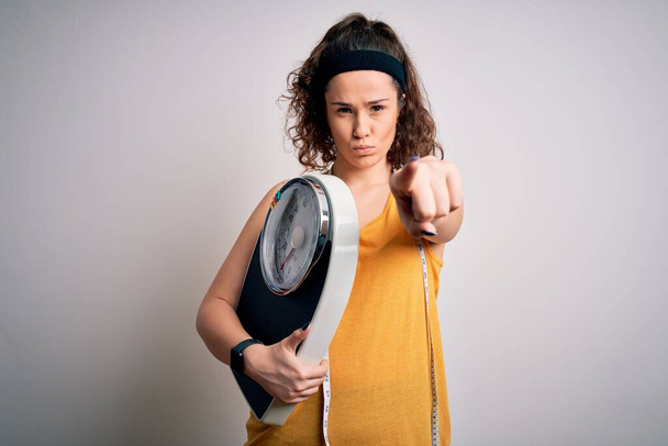 Young beautiful woman with curly hair holding weighing machine and tape measure pointing with finger to the camera and to you, hand sign, positive and confident gesture from the front - Photo, Image