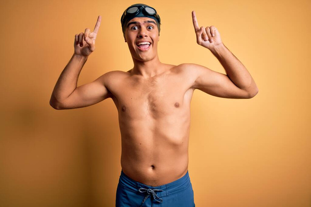 Young handsome man shirtless wearing swimsuit and swim cap over isolated yellow background smiling amazed and surprised and pointing up with fingers and raised arms. - Photo, Image