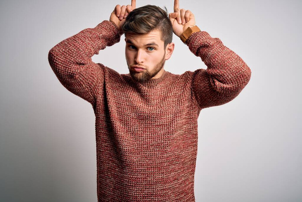 Young blond man with beard and blue eyes wearing casual sweater over white background doing funny gesture with finger over head as bull horns - Photo, Image