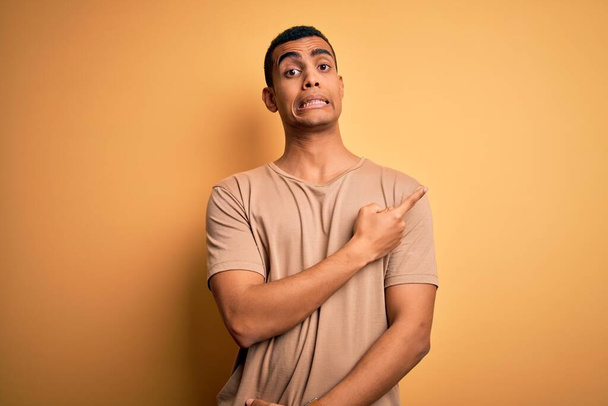 Jeune bel homme afro-américain portant un t-shirt décontracté debout sur fond jaune Pointant de côté inquiet et nerveux avec index, expression inquiète et surprise
 - Photo, image