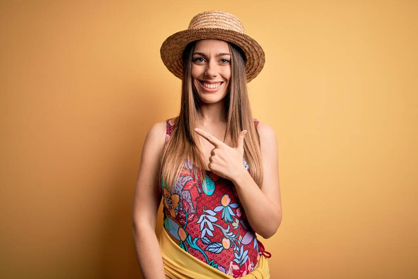 Νεαρή όμορφη ξανθιά γυναίκα με μαγιό και καλοκαιρινό καπέλο πάνω από κίτρινο φόντο χαρούμενη με χαμόγελο στο πρόσωπο δείχνοντας με το χέρι και το δάχτυλο στο πλάι με χαρούμενη και φυσική έκφραση - Φωτογραφία, εικόνα