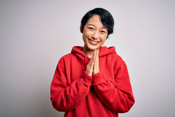 Young beautiful asian girl wearing casual sweatshirt with hoodie over white background praying with hands together asking for forgiveness smiling confident. - Fotó, kép