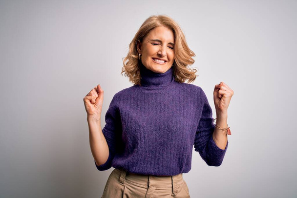 Middle age beautiful blonde woman wearing purple turtleneck sweater over white background very happy and excited doing winner gesture with arms raised, smiling and screaming for success. Celebration concept. - Photo, Image