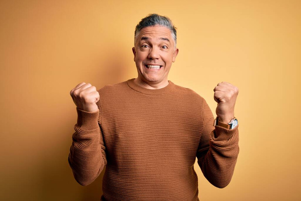 Middle age handsome grey-haired man wearing casual sweater over yellow background celebrating surprised and amazed for success with arms raised and open eyes. Winner concept. - Photo, Image