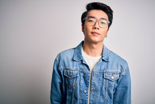 Young handsome chinese man wearing denim jacket and glasses over white background Relaxed with serious expression on face. Simple and natural looking at the camera. - 写真・画像