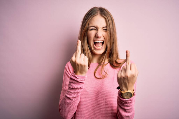 Young beautiful redhead woman wearing casual sweater over isolated pink background Showing middle finger doing fuck you bad expression, provocation and rude attitude. Screaming excited - Фото, изображение