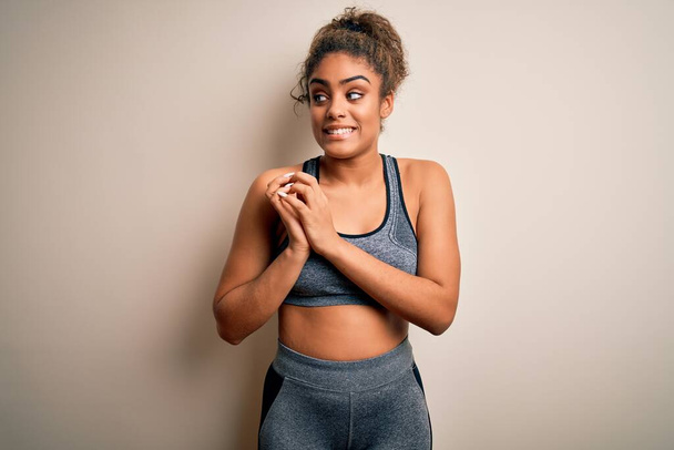 Young african american sportswoman doing sport wearing sportswear over white background laughing nervous and excited with hands on chin looking to the side - 写真・画像