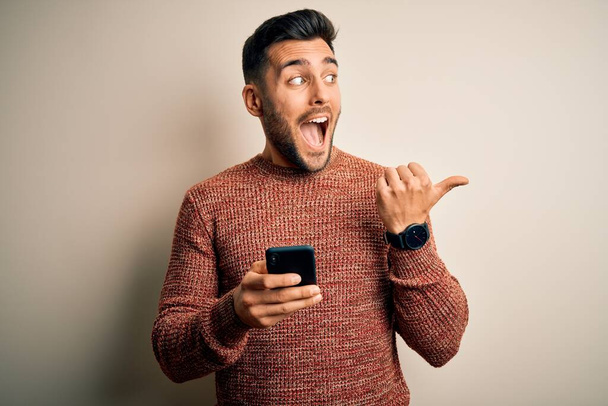 Mladý pohledný muž s rozhovorem pomocí smartphone přes bílé pozadí ukazuje palcem nahoru na stranu se šťastnou tváří usmívá - Fotografie, Obrázek