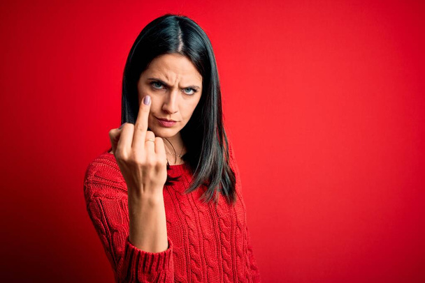 Young brunette woman with blue eyes wearing casual sweater over isolated red background Showing middle finger, impolite and rude fuck off expression - Foto, Imagen
