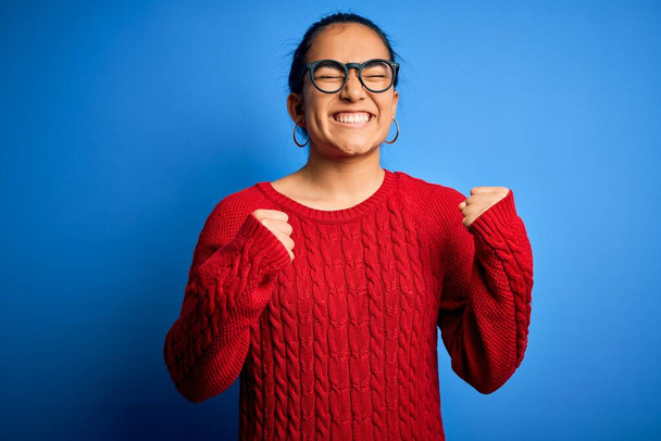 Junge schöne asiatische Frau in lässigem Pullover und Brille vor blauem Hintergrund aufgeregt für den Erfolg mit erhobenen Armen und geschlossenen Augen feiern den Sieg lächelnd. Siegerkonzept. - Foto, Bild