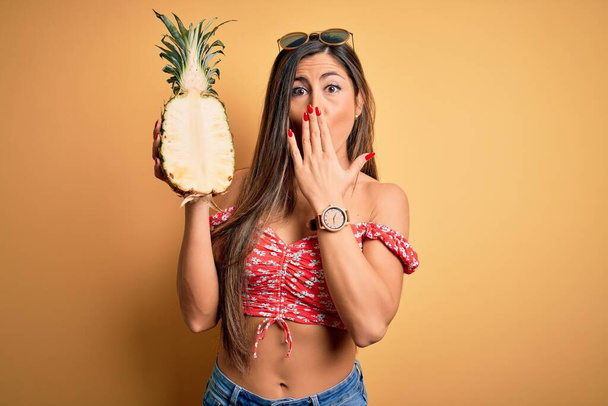 Young beautiful brunette woman holding middle pineapple fruit over isolated yellow background cover mouth with hand shocked with shame for mistake, expression of fear, scared in silence, secret concept - 写真・画像