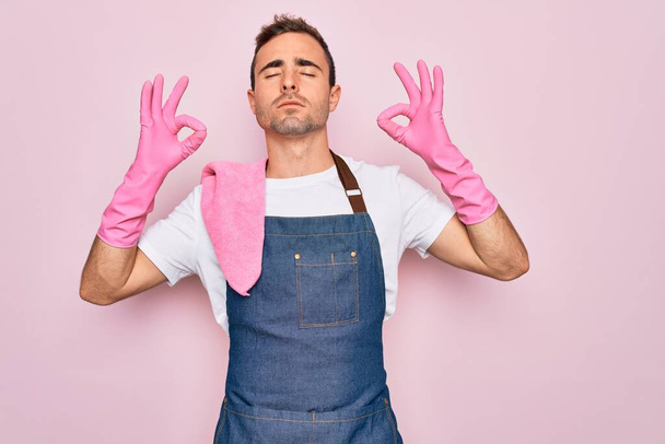 Young cleaner man with blue eyes cleaning wearing apron and gloves over pink background relax and smiling with eyes closed doing meditation gesture with fingers. Yoga concept. - Photo, Image