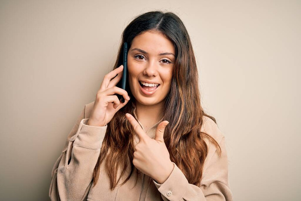Young beautiful brunette woman having conversation talking on the smartphone very happy pointing with hand and finger - 写真・画像