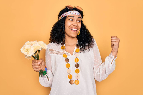 Joven mujer hippie rizada afroamericana con gafas de sol sosteniendo ramo de flores apuntando y mostrando con el pulgar hacia el lado con la cara feliz sonriendo
 - Foto, Imagen