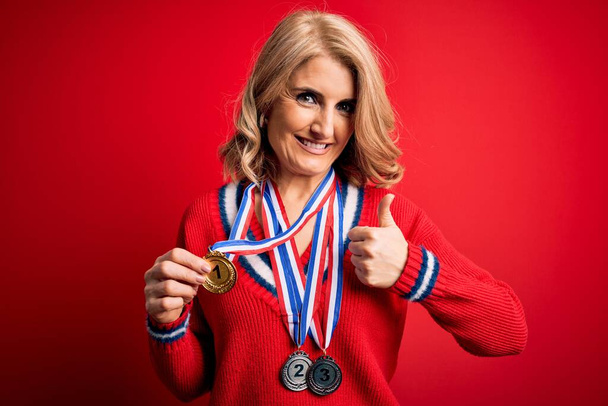 Middle age beautiful blonde successful woman winning medals over red background happy with big smile doing ok sign, thumb up with fingers, excellent sign - Photo, Image
