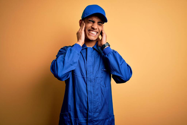 Mladý africký mechanik v modré uniformě a čepici přes žluté pozadí zakrývající uši prsty s otráveným výrazem pro hluk hlasité hudby. Hluchý koncept. - Fotografie, Obrázek