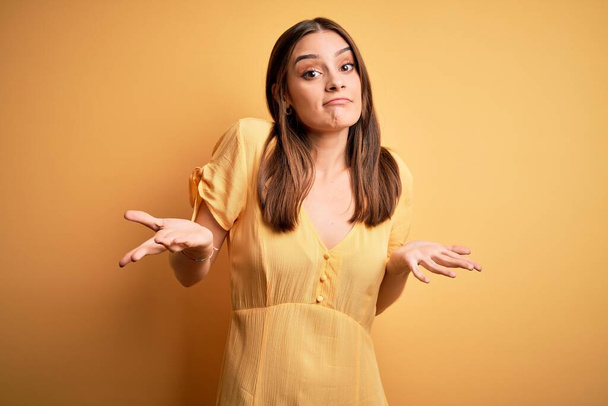 Young beautiful brunette woman wearing casual dress standing over yellow background clueless and confused expression with arms and hands raised. Doubt concept. - Photo, Image