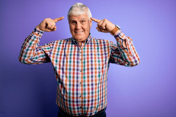 Senior pohledný chraptivý muž v ležérní barevné košili přes izolované fialové pozadí s úsměvem ukazuje na hlavu s oběma rukama prst, skvělý nápad nebo myšlenka, dobrá paměť - Fotografie, Obrázek