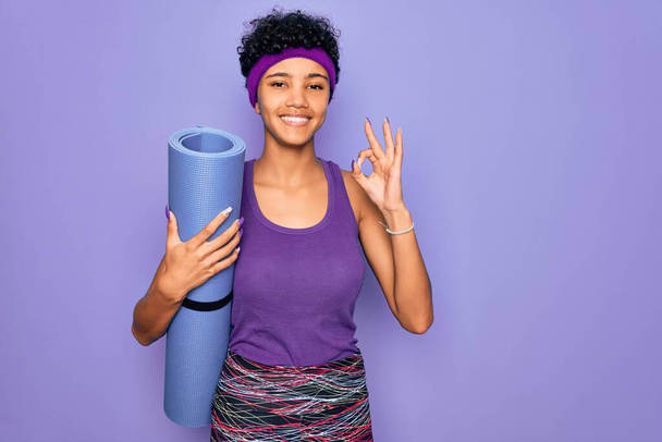 Jovem bela afro-americana desportiva mulher vestindo sportswear segurando tapete de ioga fazendo sinal ok com os dedos, excelente símbolo
 - Foto, Imagem