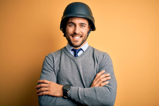 Jovem empresário bonito vestindo capacete militar sobre fundo amarelo isolado rosto feliz sorrindo com os braços cruzados olhando para a câmera. Pessoa positiva
. - Foto, Imagem