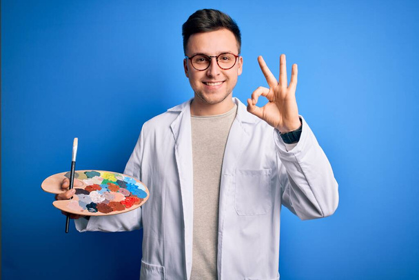 Νεαρός όμορφος καυκάσιος καλλιτέχνης άνδρας κρατώντας πινέλο και παλέτα ζωγράφος κάνει ok υπογράψει με τα δάχτυλα, εξαιρετικό σύμβολο - Φωτογραφία, εικόνα