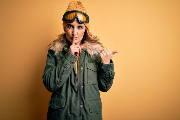 Middle age beautiful blonde skier woman wearing snow sportwear and ski goggles asking to be quiet with finger on lips pointing with hand to the side. Silence and secret concept. - Photo, Image