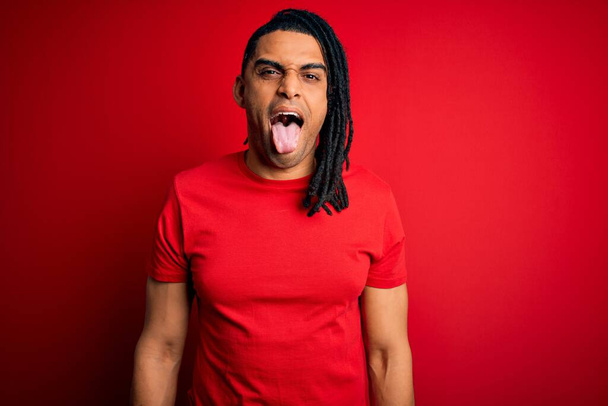 Молодий красивий афроамериканський чоловік з дредлоками в червоній повсякденній футболці, що стирчить на язику щасливим виразом. Концепція емоцій
. - Фото, зображення