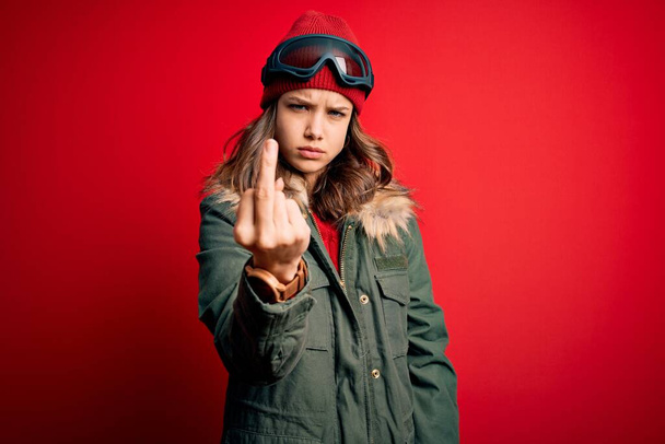 Young blonde girl wearing ski glasses and winter coat for ski weather over red background Showing middle finger, impolite and rude fuck off expression - Foto, Bild