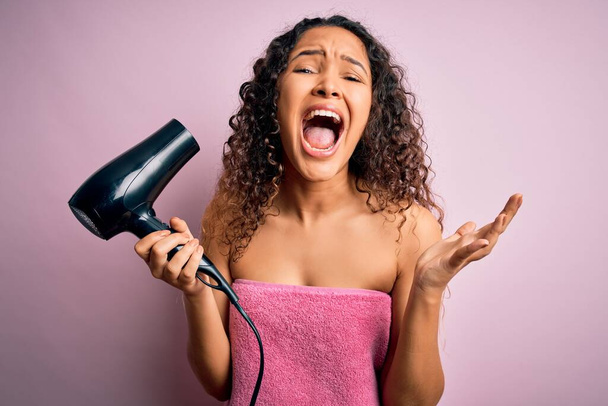 Beautiful woman with curly hair wearing shower towel after bath using hair dryer very happy and excited, winner expression celebrating victory screaming with big smile and raised hands - Photo, Image