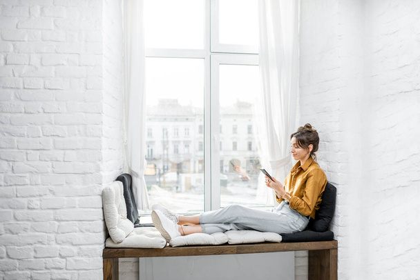 Vrouw met behulp van telefoon terwijl ontspannen op de vensterbank thuis - Foto, afbeelding